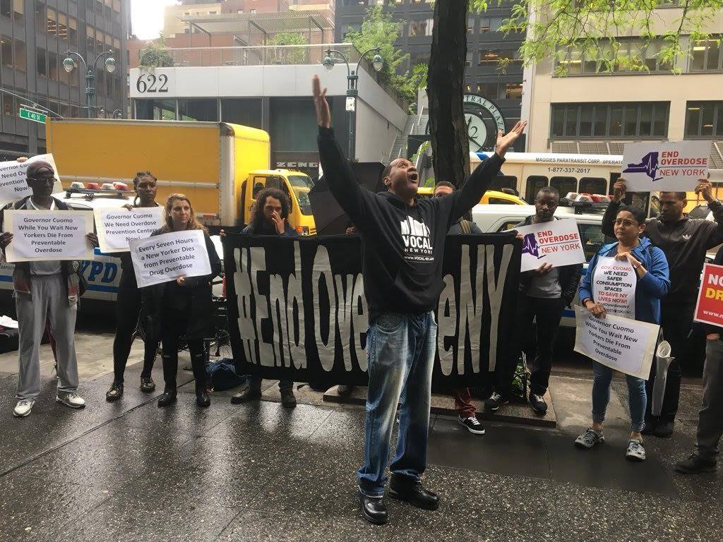 A man wearing a Vocal New York hoodie, looking up with his hands held to the sky surrounded by people holding signs and a banner that say end overdose New York