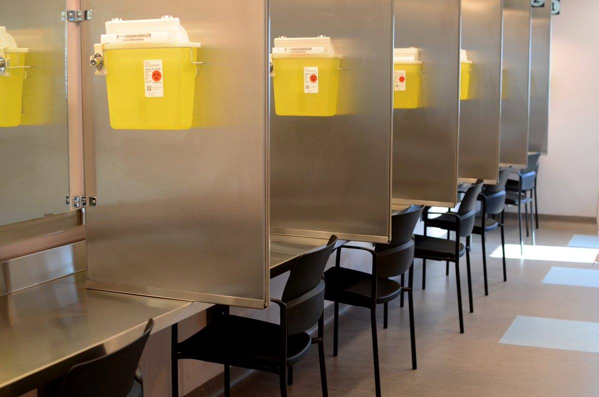 photo of a safe injection facility. A long metal table is separated by metal dividers, each with a sharps container attached to it. Chairs are put at each station.