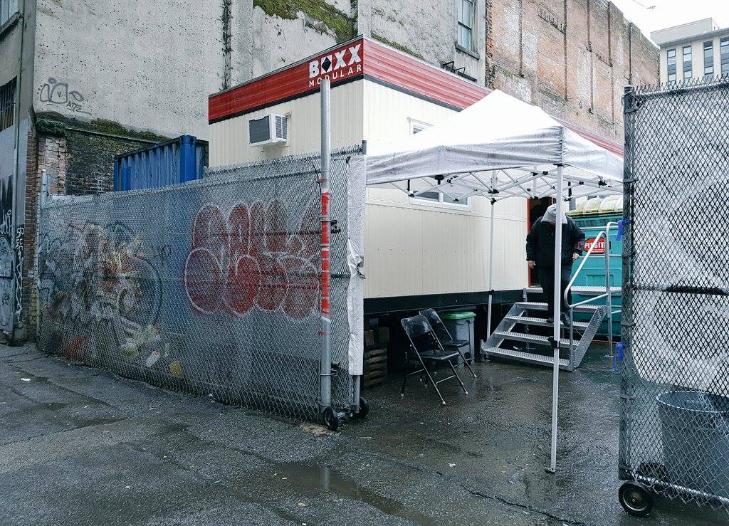 photo of the side of a building and a graffitied fence. The fence is open and inside is an easy-up tent, some chairs, and a person walking down the stairs of a modular unit that is used as a pop-up for harm reduction services.