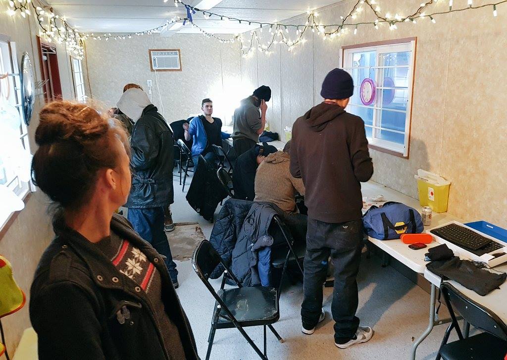 A group of people inside of a pop-up safe consumption site in Vancouver. There are tables and chairs lining the walls and string lights hanging from the ceiling.