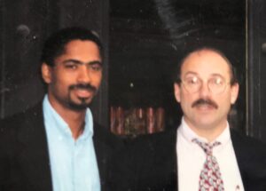 At left, Dr. Ricky Bluthenthal looks forward with a smile. He is a Black man with a mustache and beard, wearing a baby blue shirt and black suit jacket. At right is his mentor John K. Watters, a white man, wearing glasses, a white shirt with a pink patterned tie and black suit jacket.