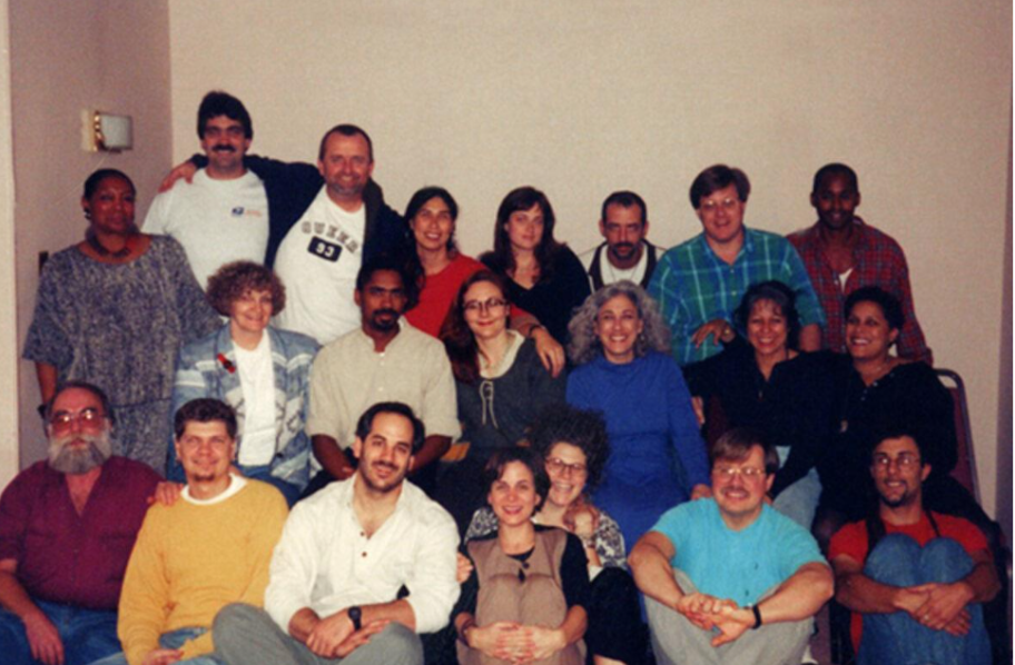 Organizing group is pictured in three rows, with folks in first two rows sitting and row in back standing. Folks are smiling, looking forward at camera view. Background is cream/pink colored interior walls.