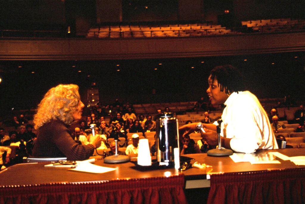 Imani Woods at right, a Black woman wearing white, speaking with another person at a table on auditorium stage. In background, audience sitting in auditorium seats listens on.