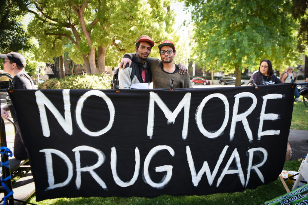 Image of two people standing behind large banner reading, "NO MORE DRUG WAR". They have their arms around each other's shoulders, and are looking directly at camera.