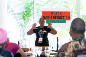 In focus at center, Black woman wearing lanyard, black shirt, name tag, and glasses, speaks to audience, gesturing with her hands. Behind her is sign reading, "BLACK HARM REDUCTION Network". In foreground out of focus are audience members listening in. 